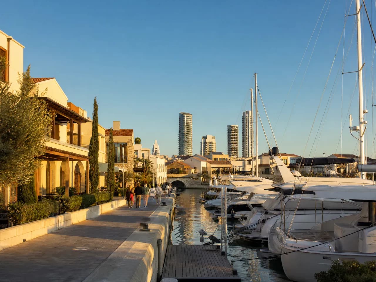 Marina with white yachts along a quay, low stone buildings and towers in the distance. Illustrative image, not a photograph of a specific place.