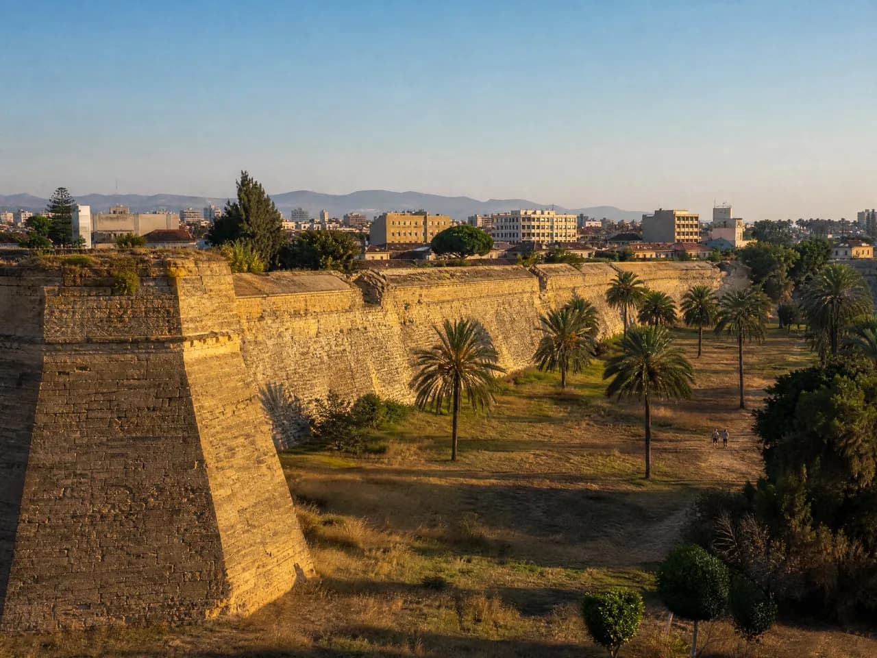 Sandstone bastion wall with palm trees in the dry moat below it and low city buildings behind. Illustrative image, not a photograph of a specific place.